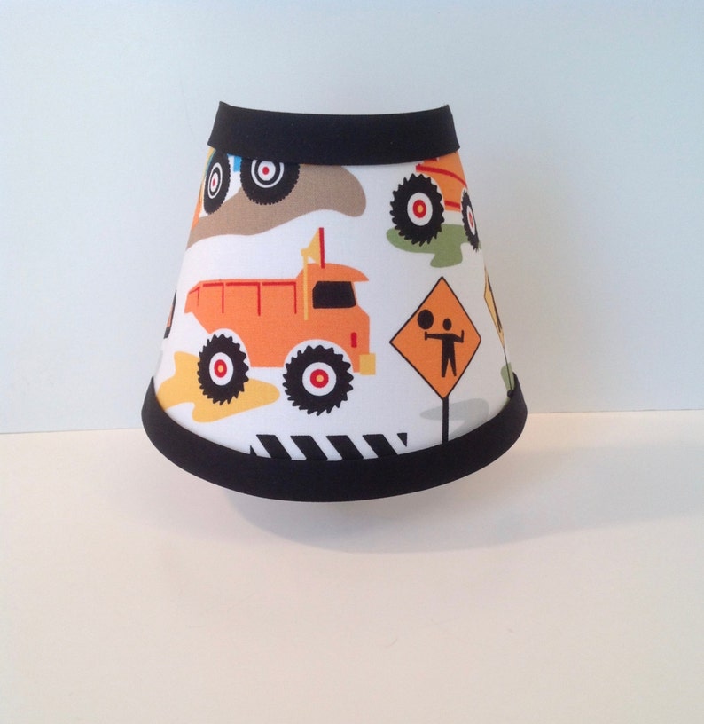 May include: A white lampshade with a black trim features a construction theme with orange dump trucks, black and white striped tires, and a yellow road sign with a stick figure.