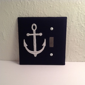 May include: Navy blue light switch cover with a white embroidered anchor design. The nautical-themed cover has a rectangular shape with a toggle switch and two screw holes. A decorative accent for a coastal or nautical decor.