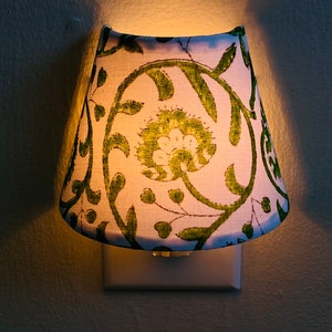 Puede incluir: Una luz nocturna blanca con una pantalla de estampado floral verde. La pantalla est&aacute; enchufada a una toma de corriente est&aacute;ndar.