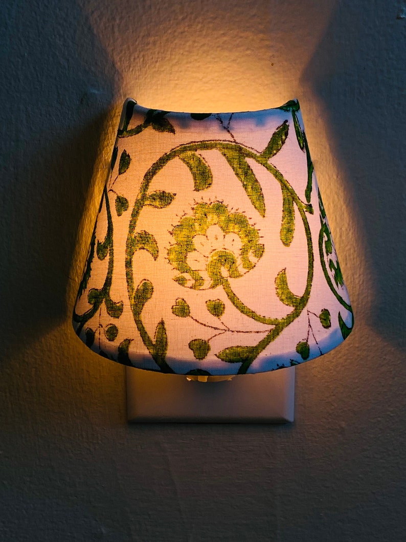 Puede incluir: Una luz nocturna blanca con una pantalla de estampado floral verde. La pantalla est&aacute; enchufada a una toma de corriente est&aacute;ndar.
