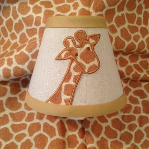 May include: A beige lampshade with a brown giraffe applique. The lampshade has a gold trim around the top and bottom.