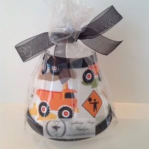 May include: A white lampshade with a black trim and a construction vehicle design. The lampshade is wrapped in clear plastic and tied with a black ribbon. The label on the lampshade reads "Lightning Bugs Boutique" and "www.LightningBugsEtsy.com".