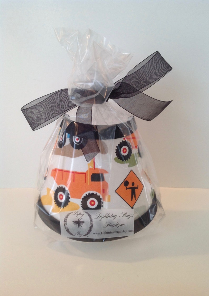 May include: A white lampshade with a black trim and a construction vehicle design. The lampshade is wrapped in clear plastic and tied with a black ribbon. The label on the lampshade reads "Lightning Bugs Boutique" and "www.LightningBugsEtsy.com".
