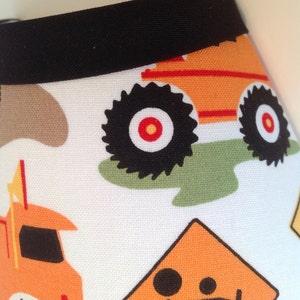 May include: White fabric with a black trim and a repeating pattern of orange construction vehicles. The vehicles have black wheels with red centers. There is also a yellow construction sign with a black figure.