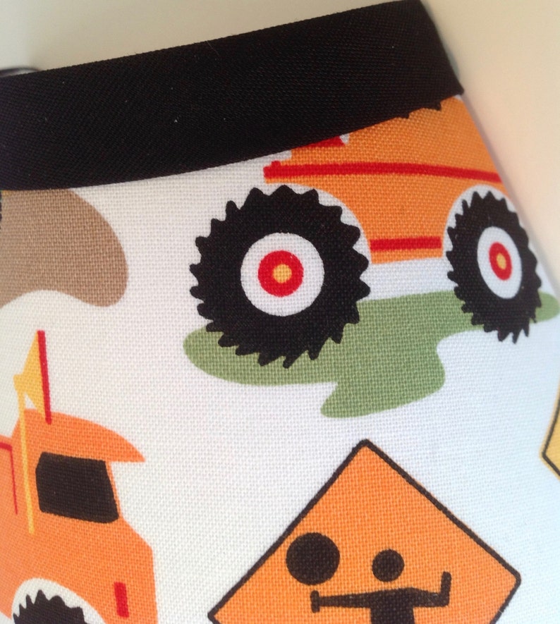 May include: White fabric with a black trim and a repeating pattern of orange construction vehicles. The vehicles have black wheels with red centers. There is also a yellow construction sign with a black figure.