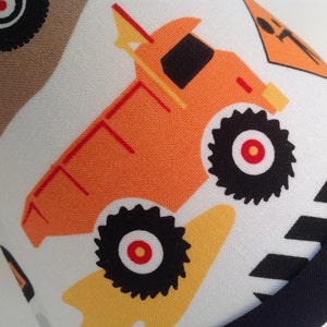 May include: A white fabric with a black and white striped border. The fabric features a repeating pattern of orange dump trucks with black wheels and red centers. The trucks have yellow accents.