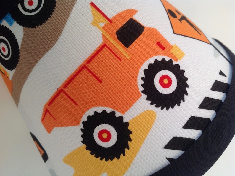 May include: A white fabric with a black and white striped border. The fabric features a repeating pattern of orange dump trucks with black wheels and red centers. The trucks have yellow accents.