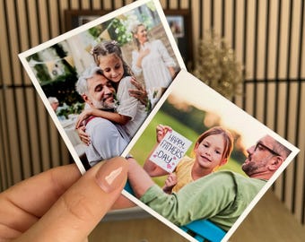 May include: Two square photographs are held in a hand. One shows a girl hugging a man, the other shows a girl holding a sign that says "Happy Father's Day." The photos are on a wooden background.
