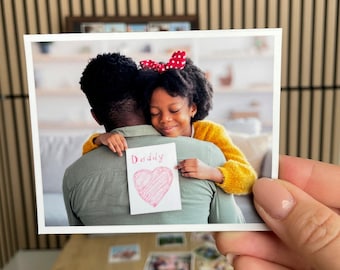 May include: A photograph of a young girl hugging a person, holding a card that says "Daddy" with a red heart. The girl is wearing a yellow sweater and a red polka dot bow. The photo is held by a hand.