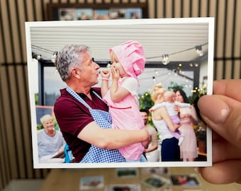 May include: A photograph of a family gathering. An older man in a maroon shirt and blue apron holds a young child in a pink and white striped outfit and bonnet. Other family members are visible in the background.
