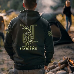 May include: A dark green hooded sweatshirt with a graphic of a coiled snake over an American flag design. The text "DON'T TREAD ON THEIR SACRIFICE" is printed on the back. The sweatshirt has a hood and a kangaroo pocket.