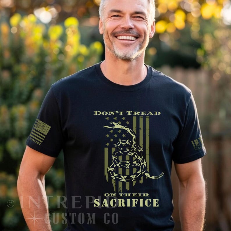 May include: Black t-shirt featuring a distressed American flag design with a coiled snake and the words "DON'T TREAD" and "ON THEIR SACRIFICE". The shirt also has a small American flag on the left sleeve and the word "TRUST".