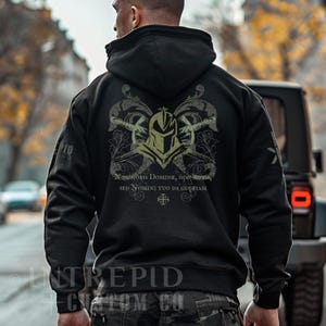 May include: Black hoodie with a hood and a graphic design on the back. The design features a helmet, crossed weapons, and text in a decorative style. The text includes the phrases "NON NOBIS DOMINE, NON NOBIS, SED NOMINI TUO DA GLORIAM" and "INTREPID CUSTOM CO."