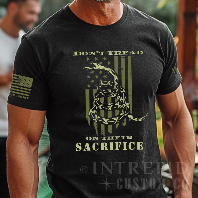 May include: Black t-shirt featuring a coiled snake and the text "DON'T TREAD ON THEIR SACRIFICE." The design includes an American flag with stars and stripes, and the words "WE TRUST" on the sleeve.