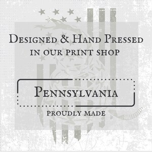 May include: A light gray graphic with the text "Designed & Hand Pressed in Our Print Shop" above a rectangular box with the word "Pennsylvania" inside. The words "Proudly Made" are below the box. The background has a distressed look.