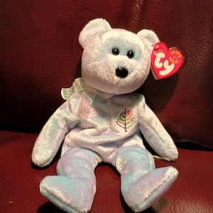 Puede incluir: Un oso de peluche Beanie Baby azul claro con una etiqueta Ty roja. El oso tiene un pequeño árbol bordado en el pecho y una cinta blanca alrededor del cuello.