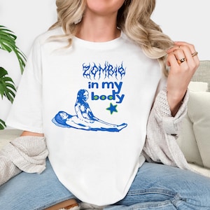 May include: A white short-sleeve t-shirt featuring a blue graphic print. The design includes the text "ZOMBIE in my body" in a stylized font, a blue star with a green outline, and an illustration of one figure lying down while another sits.
