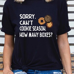 May include: Black t-shirt with the text "SORRY. CAN'T COOKIE SEASON. HOW MANY BOXES?" and illustrations of cookies. The shirt is paired with blue jeans. The design is perfect for cookie lovers.