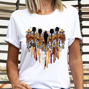 May include: White t-shirt featuring a colorful, abstract design of feathers with dripping paint effect. The design incorporates shades of brown, yellow, and red. The shirt is a casual wear item.