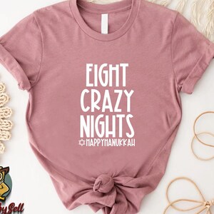 May include: A dusty rose t-shirt with the text "EIGHT CRAZY NIGHTS" in white, with "HAPPY HANUKKAH" below. The shirt is knotted at the bottom. The design is perfect for celebrating Hanukkah.