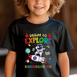 May include: Black short-sleeved t-shirt with the text "Ready to Explore Kindergarten." The graphic features an astronaut on a rocket, a globe, books, and an apple. The design is in bright, colorful letters.