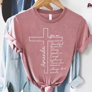 May include: A dusty rose t-shirt with a white cross design. The cross includes the text "You are Beautiful, Strong, Enough, Never alone, Always Loved" and the name "Amanda". The shirt is hanging on a wooden hanger.