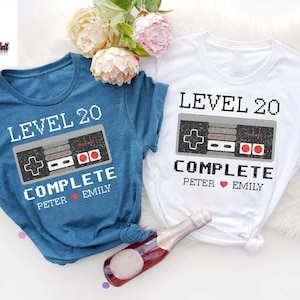 May include: Two blue and white t-shirts with a retro video game controller graphic and the text "Level 20 Complete" with names Peter and Emily.