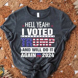 May include: A dark gray t-shirt with the text "HELL YEAH! I VOTED TRUMP AND WILL DO IT AGAIN 2024" printed in white. The text is surrounded by stars and the word "TRUMP" is printed in red, white, and blue.
