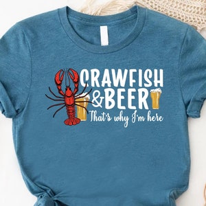 May include: A teal blue t-shirt with a white graphic of a crawfish holding a beer glass. The text reads "Crawfish & Beer That's why I'm here."