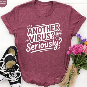 May include: Heather maroon t-shirt with the text "ANOTHER VIRUS? Seriously?" in white. The shirt is paired with black and white sneakers and a bouquet of pink, purple, and orange flowers.