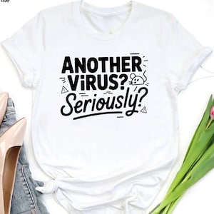 May include: White t-shirt with the text "ANOTHER VIRUS? Seriously?" in black. The design includes a mouse and geometric shapes. The shirt is short-sleeved and appears to be made of cotton.