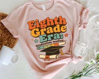 May include: A light pink short-sleeve t-shirt featuring the text "Eighth Grade Era: Graduation loading..." in a retro font. The graphic includes a black graduation cap with a yellow tassel, a rolled diploma, and a colourful horizontal bar.