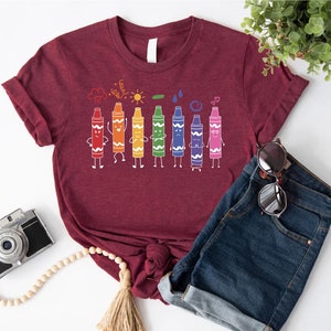 May include: A maroon t-shirt with a graphic of colorful crayons with faces and arms. The crayons are lined up in a row and are drawing a cloud, sun, raindrop, circle, and heart.