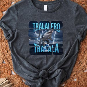 May include: A dark gray t-shirt with a graphic of a shark jumping out of the water with lightning bolts and the text "TRALAIRO TRALALA".