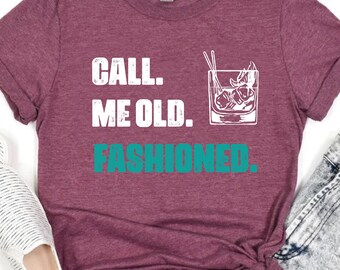 May include: A maroon t-shirt with the text "CALL. ME OLD. FASHIONED." in white and teal. A graphic of a cocktail is next to the text. The shirt is paired with denim shorts and white slip-on shoes.