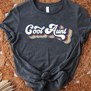 May include: Dark gray t-shirt with the words "Cool Aunt" in a retro font with blue, orange, and cream colors. The shirt is knotted at the bottom and is displayed on a woven, circular rug.
