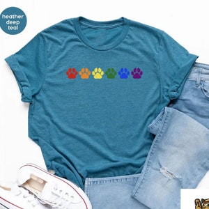 May include: A teal blue t-shirt with a row of rainbow paw prints. The shirt is laid flat on a white surface with a pair of white sneakers and light blue jeans.