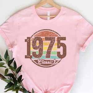 May include: A light pink t-shirt with a brown circular graphic that reads "1975 Classic" in a retro style. The graphic features a rainbow-colored stripe design. The shirt is hanging on a wooden hanger.