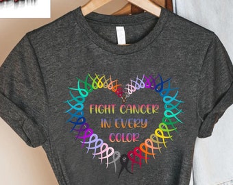 Puede incluir: Camiseta gris oscuro con un diseño en forma de corazón hecho de cintas coloridas. El texto "FIGHT CANCER IN EVERY COLOR" está centrado dentro del corazón. La camiseta está colgada en una percha de madera.
