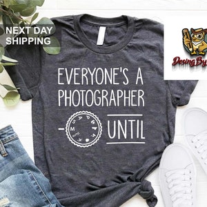 May include: Gray t-shirt with the text "Everyone's a Photographer Until" and a camera aperture setting graphic. The shirt is displayed on a white surface with a pair of white sneakers.