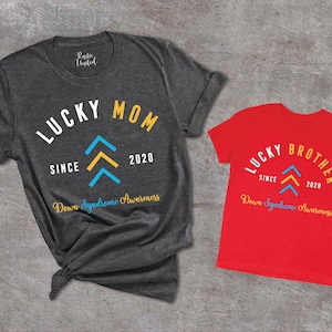 Op de afbeelding: Twee T-shirts, een grijs en een rood, met de tekst "Lucky Mom Since 2020 Down Syndrome Awareness" en "Lucky Brother Since 2020 Down Syndrome Awareness". Beide shirts hebben een gestileerd geel en blauw pijlontwerp.