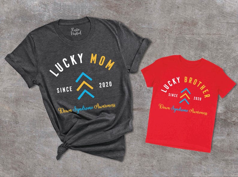 Op de afbeelding: Twee T-shirts, een grijs en een rood, met de tekst "Lucky Mom Since 2020 Down Syndrome Awareness" en "Lucky Brother Since 2020 Down Syndrome Awareness". Beide shirts hebben een gestileerd geel en blauw pijlontwerp.