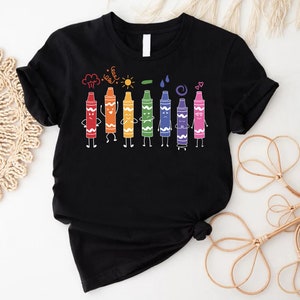 May include: Black t-shirt with a rainbow of crayons with faces and arms. The crayons are lined up and have different expressions. Above the crayons are drawings of a cloud, sun, leaf, raindrops, and a swirl.