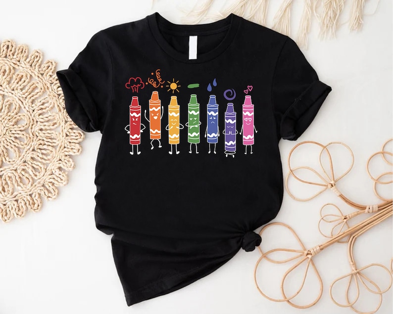 May include: Black t-shirt with a rainbow of crayons with faces and arms. The crayons are lined up and have different expressions. Above the crayons are drawings of a cloud, sun, leaf, raindrops, and a swirl.