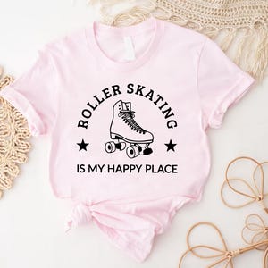 May include: Light pink t-shirt with a black graphic of a roller skate and the text "ROLLER SKATING IS MY HAPPY PLACE". The design includes two black stars. The shirt is made of a soft material.