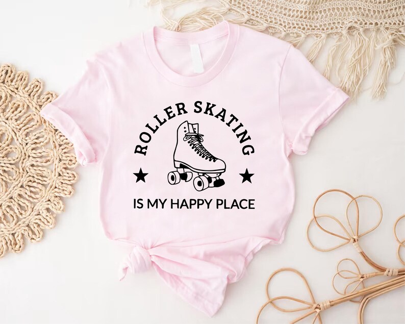 May include: Light pink t-shirt with a black graphic of a roller skate and the text "ROLLER SKATING IS MY HAPPY PLACE". The design includes two black stars. The shirt is made of a soft material.
