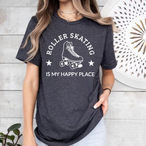 May include: Dark gray t-shirt with the text "ROLLER SKATING IS MY HAPPY PLACE" in white. The design features a roller skate illustration with stars. The shirt is a crew neck and has short sleeves.