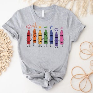 May include: Gray t-shirt with a rainbow of crayons with faces and arms. The crayons are arranged in a line and are smiling, frowning, and making other expressions. The text "Cray-Z" is above the crayons.