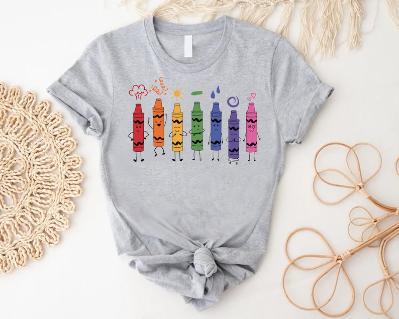 May include: Gray t-shirt with a rainbow of crayons with faces and arms. The crayons are arranged in a line and are smiling, frowning, and making other expressions. The text "Cray-Z" is above the crayons.