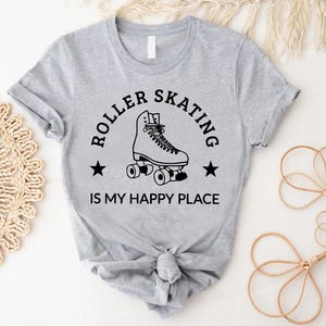 May include: A light gray t-shirt with a black graphic that says "ROLLER SKATING IS MY HAPPY PLACE." The graphic features a roller skate and two stars. The shirt is tied in a knot at the bottom.